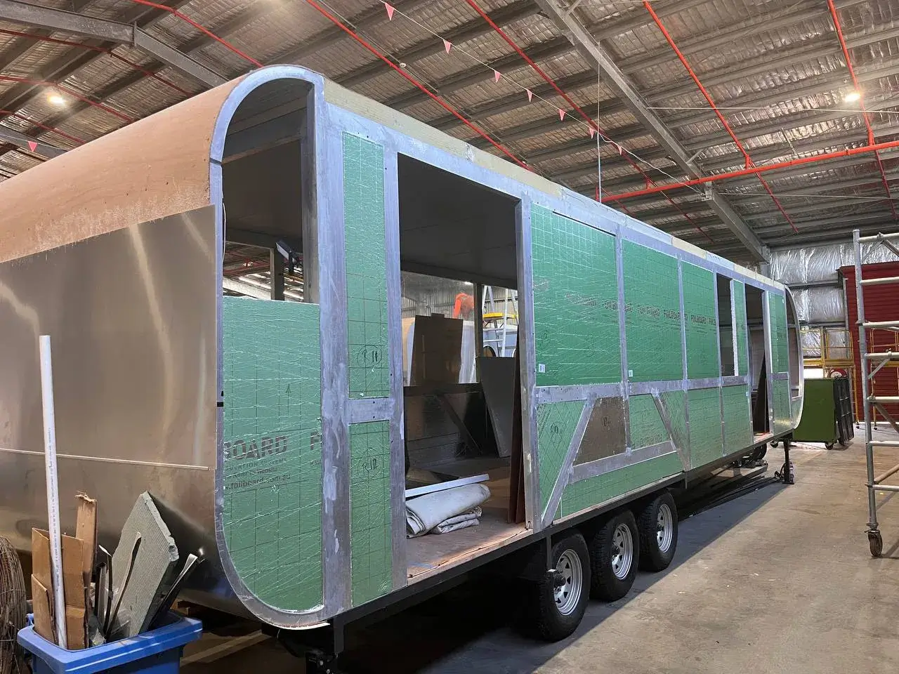 Side view of a 37ft long caravan under construction, showing green insulation panels and an aluminum frame in a workshop.